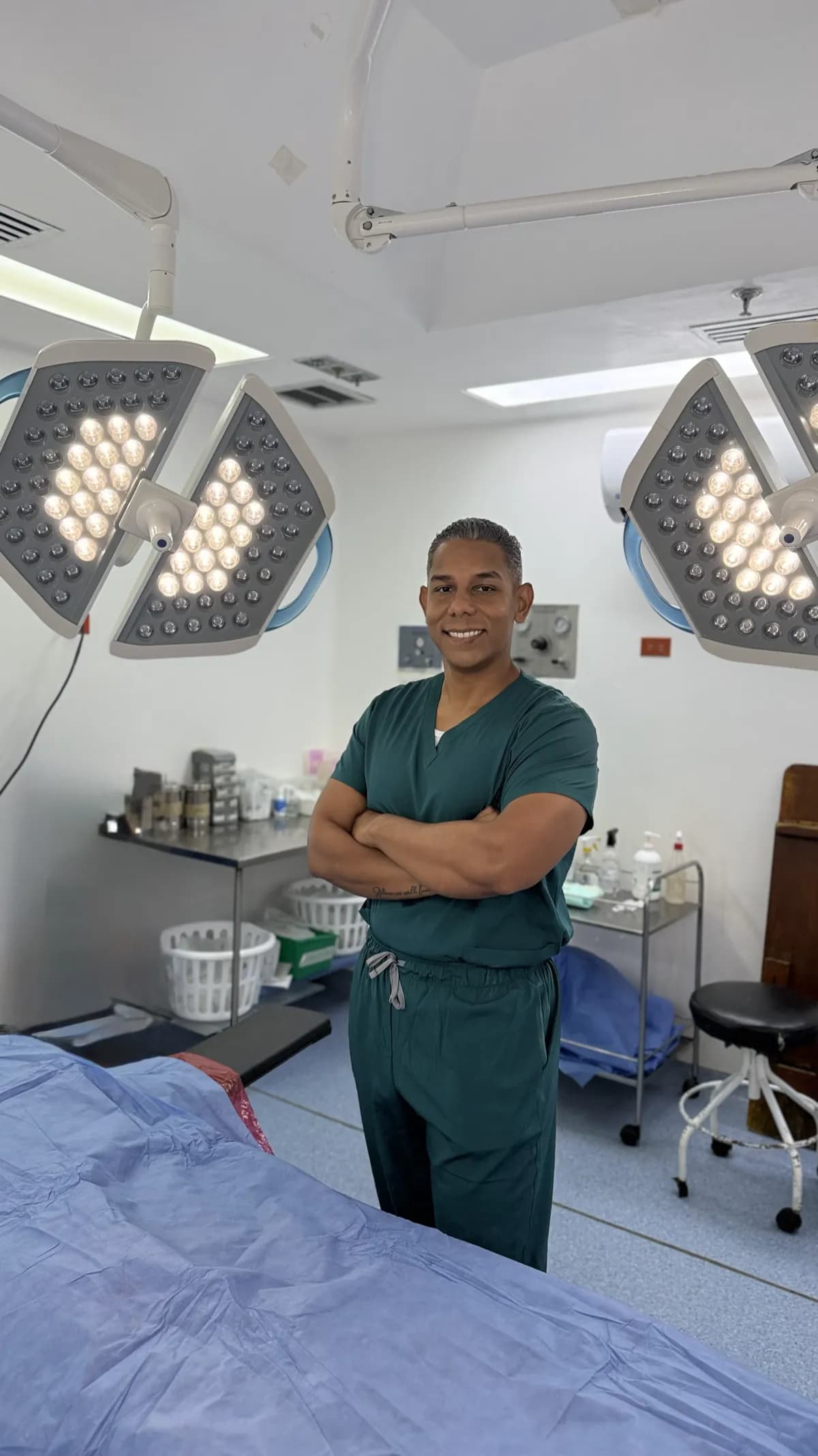El Dr. Camelo sonríe de brazos cruzados en el quirófano, bajo las lámparas encendidas.
